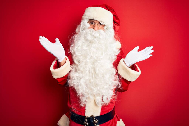 Ein gutaussehender Mann mittleren Alters im Weihnachtsmannkostüm steht ratlos und verwirrt mit erhobenen Armen und Händen vor isoliertem roten Hintergrund. Zweifelhaftes Konzept. - Foto, Bild