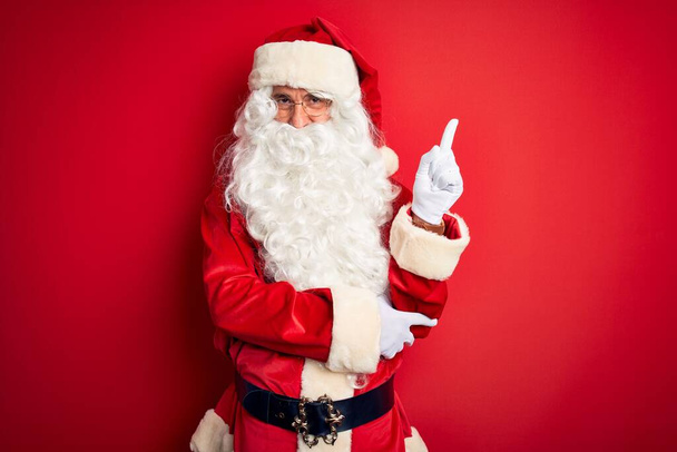 Hombre guapo de mediana edad vistiendo traje de Santa de pie sobre un fondo rojo aislado con una gran sonrisa en la cara, señalando con el dedo de la mano hacia el lado mirando a la cámara
. - Foto, Imagen