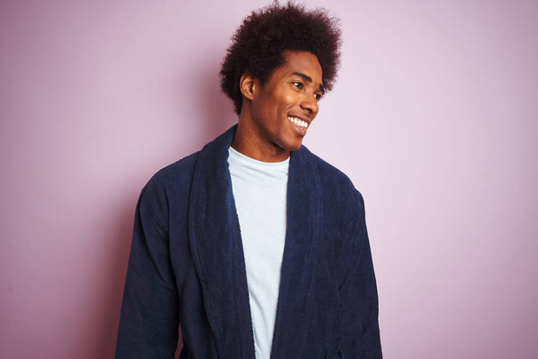 Fiatal afro-amerikai férfi pizsamát visel, elszigetelt rózsaszín háttér fölött áll, elfordul a mosollyal az arcán, természetes kifejezéssel. Nevetve magabiztos. - Fotó, kép
