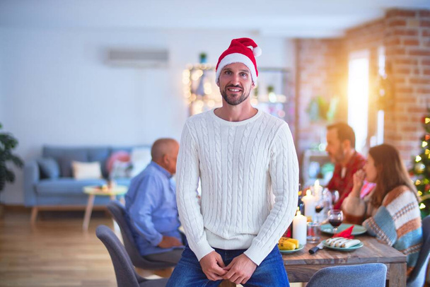 Jovem bonitão sorrindo feliz e confiante. De pé usando chapéu de Papai Noel celebrando o Natal com a família em casa
 - Foto, Imagem