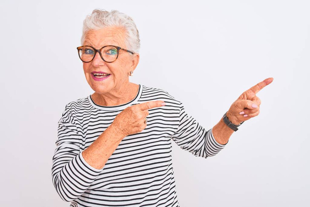 Senior harmaa tukka nainen yllään raidallinen laivaston t-paita lasit yli eristetty valkoinen tausta hymyillen ja katsoen kameraa osoittaa kahdella kädellä ja sormella sivulle
. - Valokuva, kuva