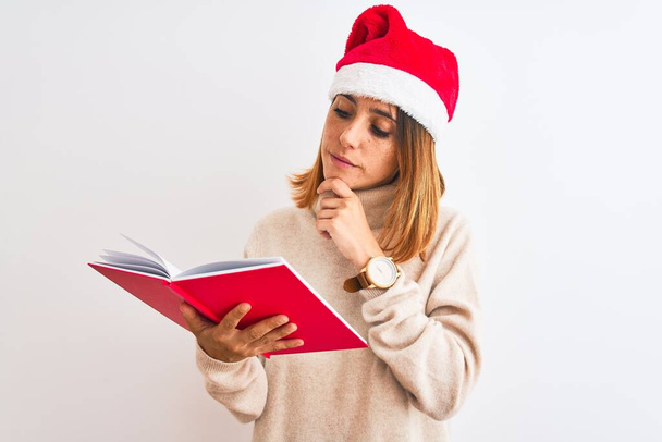 Gyönyörű vörös hajú nő visel karácsonyi kalap és olvasás egy könyvet elszigetelt háttér komoly arc gondolkodás kérdés, nagyon zavaros ötlet - Fotó, kép