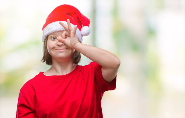 Mulher adulta jovem com síndrome de down usando chapéu de Natal sobre fundo isolado fazendo gesto ok com a mão sorrindo, olho olhando através dos dedos com rosto feliz
. - Foto, Imagem