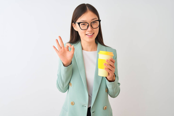 Młoda Chinka w okularach pije kawę na białym tle robi znak ok palcami, doskonały symbol - Zdjęcie, obraz