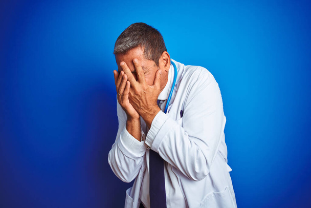 Красивый врач средних лет носит стетоскоп на изолированном синем фоне с грустным лицом, прикрывающим лицо руками во время плача. Концепция депрессии
. - Фото, изображение