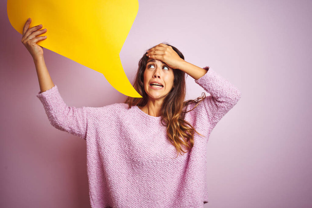 Young beautiful woman holding yellow speech bubble standing over pink isolated background stressed with hand on head, shocked with shame and surprise face, angry and frustrated. Fear and upset for mistake. - Photo, Image