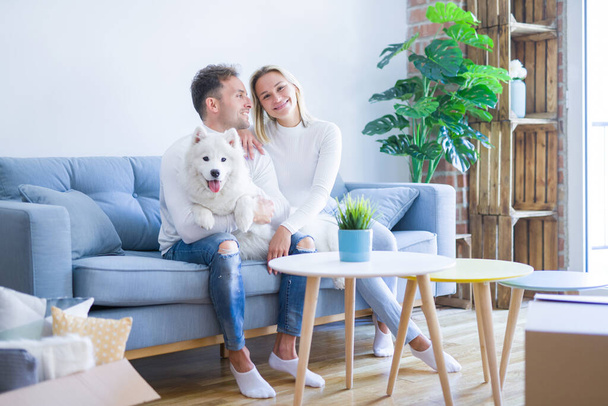 Young beautiful couple with dog sitting on the sofa at new home around cardboard boxes - Fotó, kép