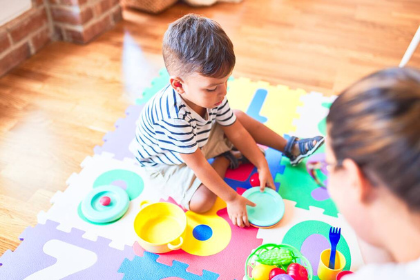Hermoso niño sentado en el rompecabezas jugando con platos de plástico, frutas y verduras en el jardín de infantes
 - Foto, Imagen