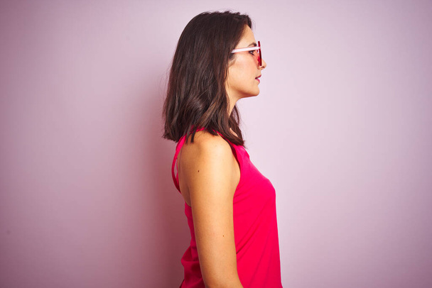 Красива жінка в смішних рожевих окулярах у формі серця над рожевим ізольованим тлом, дивлячись в бік, розслабляє профільну позу з природним обличчям з впевненою посмішкою
. - Фото, зображення