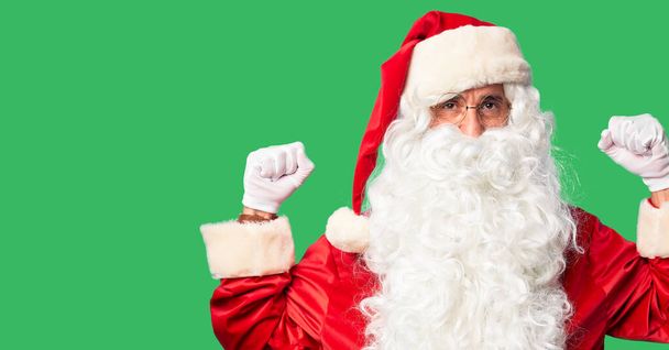 Красивый мужчина средних лет в костюме Санта-Клауса и с бородой на руках, гордо улыбающийся мускулами. Фитнес-концепция
. - Фото, изображение