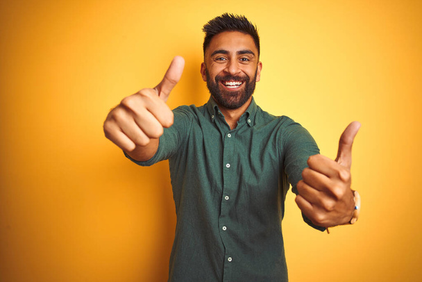 Молодий індійський бізнесмен в елегантній сорочці, що стоїть над ізольованим білим тлом, схвалюючи позитивний жест рукою, великі пальці посміхаються і щасливі за успіх. жест переможця
. - Фото, зображення