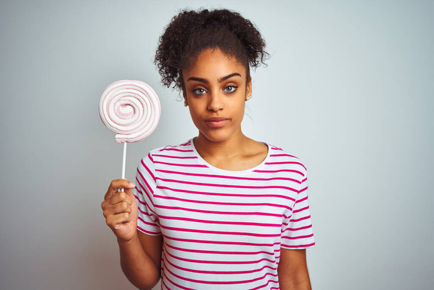 Afro-Amerikalı genç bir kadın izole edilmiş beyaz arka planda renkli şekerler yiyor. Akıllı bir yüz ifadesiyle ciddi ciddi düşünüyor. - Fotoğraf, Görsel