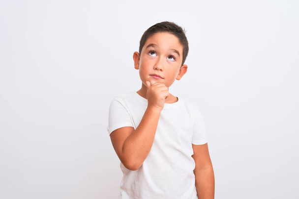 Beautiful kid boy wearing casual t-shirt standing over isolated white background with hand on chin thinking about question, pensive expression. Smiling with thoughtful face. Doubt concept. - Photo, Image