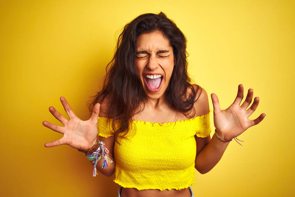 Young beautiful woman wearing casual t-shirt standing over isolated yellow background celebrating mad and crazy for success with arms raised and closed eyes screaming excited. Winner concept - Fotografie, Obrázek