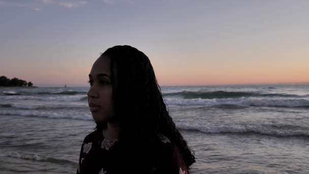 Handheld slow motion shot krążący wokół głowy spokojnej młodej Afroamerykanki mieszanej rasy kobiety stojącej na piaszczystej plaży, gdy fale toczą się za nią o wschodzie słońca.. - Materiał filmowy, wideo