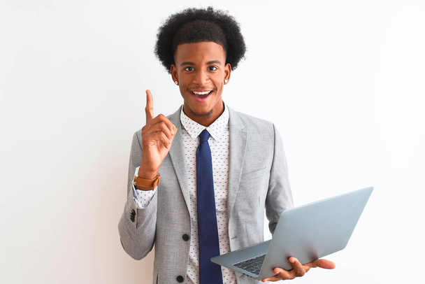 Молодой африканский американский бизнесмен, использующий ноутбук, стоящий на изолированном белом фоне, удивлен идеей или вопросом, указывая пальцем со счастливым лицом, номер один
 - Фото, изображение