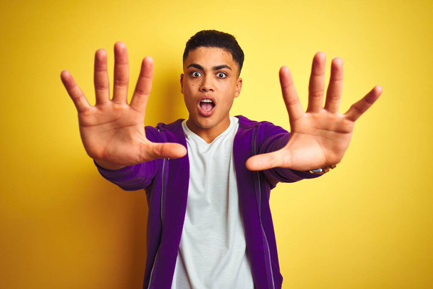Young brazilian man wearing purple sweatshirt standing over isolated yellow background doing stop gesture with hands palms, angry and frustration expression - 写真・画像