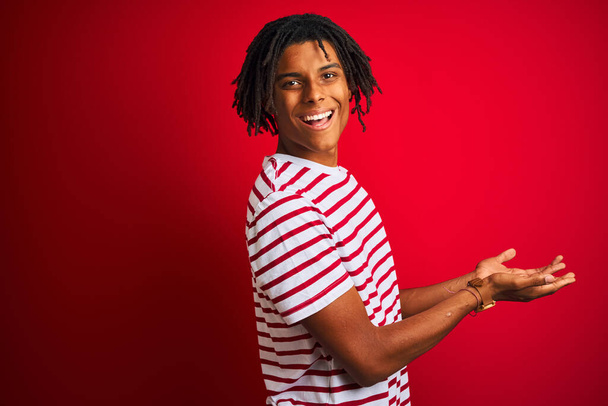 jovem afro homem com dreadlocks vestindo listrado t-shirt de pé sobre isolado fundo vermelho apontando para o lado com as mãos abertas palmas mostrando espaço cópia, apresentando anúncio sorrindo feliz animado
 - Foto, Imagem