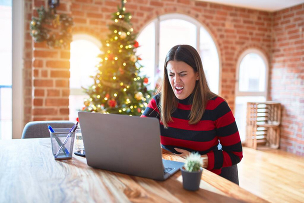 Mooie vrouw zit aan tafel te werken met laptop thuis rond kerstboom met hand op buik omdat misselijkheid, pijnlijke ziekte zich niet goed voelt. Ache-concept. - Foto, afbeelding