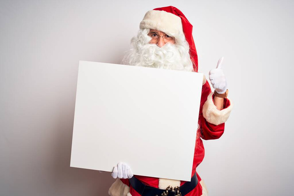 Middle age man wearing Santa Claus costume holding banner over isolated white background happy with big smile doing ok sign, thumb up with fingers, excellent sign - Foto, afbeelding