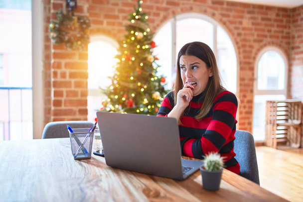 Belle femme assise à la table travaillant avec un ordinateur portable à la maison autour de l'arbre de Noël regardant stressé et nerveux avec les mains sur la bouche mordant ongles. Problème d'anxiété
. - Photo, image
