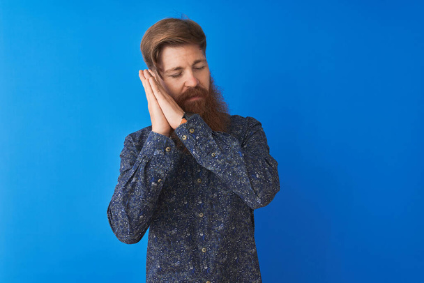 Young redhead irish man wearing floral summer shirt standing over isolated blue background sleeping tired dreaming and posing with hands together while smiling with closed eyes. - Photo, Image