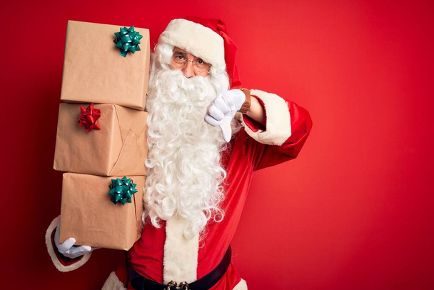 Homem sênior vestindo traje de Papai Noel segurando torre de presentes sobre fundo vermelho isolado com rosto irritado, sinal negativo mostrando antipatia com os polegares para baixo, conceito de rejeição
 - Foto, Imagem