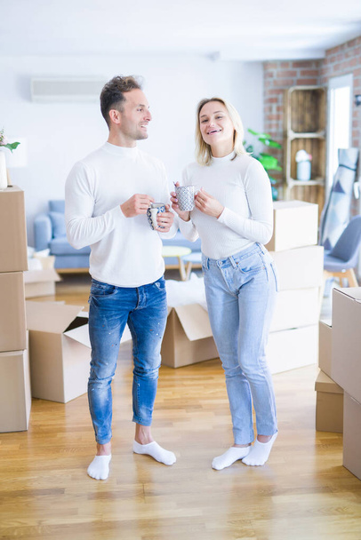 Young beautiful couple standing drinking cup of coffee at new home around cardboard boxes - Фото, зображення