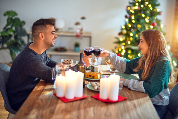 Jovem casal bonito sorrindo feliz e confiante. Comer alimentos e brindar com xícara de vinho celebrando o Natal em casa
 - Foto, Imagem