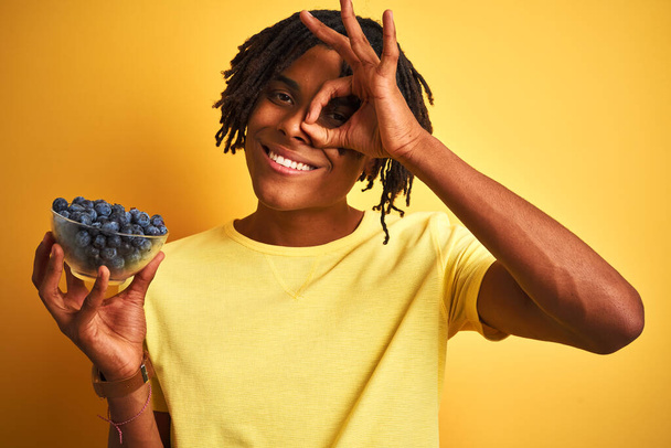 Afro amerykański człowiek z dredami trzymając jagody na odizolowanym żółtym tle z uśmiechem szczęśliwej twarzy robi ok znak z ręki na oko patrząc przez palce - Zdjęcie, obraz