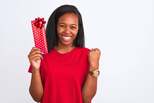 Jeune femme afro-américaine tenant cadeau d'anniversaire sur fond isolé criant fier et célébrant la victoire et le succès très excité, acclamant l'émotion
 - Photo, image
