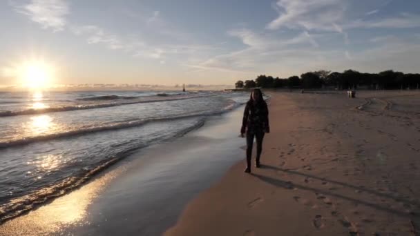 A beautiful African American woman walks along the smooth sand in jeans and long sleeve shirt as the sun on the horizon reflects over the crashing waves along the shoreline Lake Michigan in Chicago. - Materiał filmowy, wideo