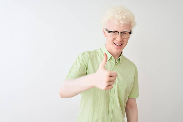 junger Albino-blonder Mann mit grünem Hemd und Brille vor isoliertem weißem Hintergrund, der fröhlich mit der Hand Daumen nach oben gestikuliert. zustimmender Ausdruck, der mit Erfolg in die Kamera blickt. - Foto, Bild