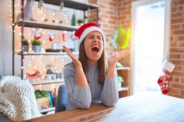 Молода красива жінка в різдвяному капелюсі сидить за столом вдома божевільно і божевільно кричить і кричить з агресивним виразом і піднятими руками. Концепція розмальовки
. - Фото, зображення
