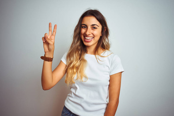 Giovane bella donna indossa casual t-shirt bianca su sfondo isolato mostrando e indicando con le dita numero due, sorridendo fiducioso e felice
. - Foto, immagini