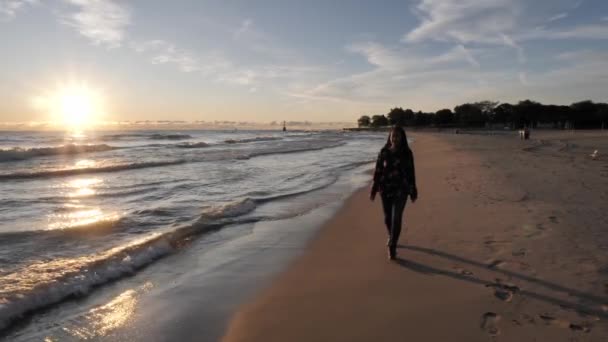 A beautiful African American woman walks along the smooth sand in jeans and long sleeve shirt as the sun on the horizon reflects over the crashing waves along the shoreline Lake Michigan in Chicago. - Záběry, video