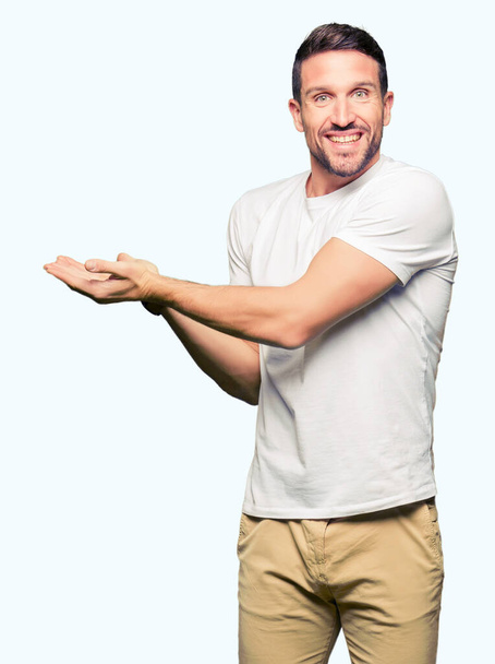 Красивий чоловік у повсякденній білій футболці вказує на бік рукою і відкритою долонею, представляючи рекламу щасливою і впевненою
 - Фото, зображення