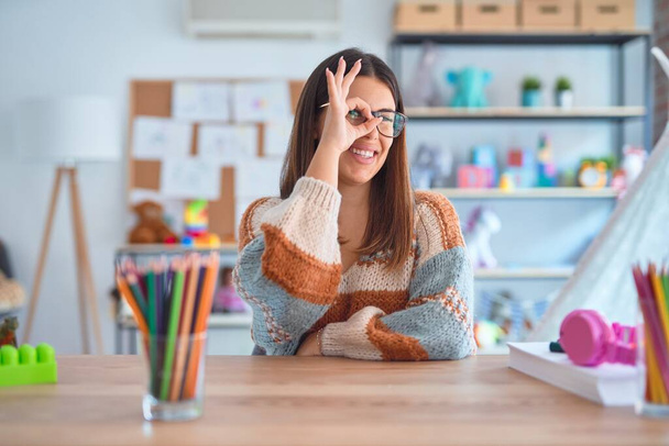 Joven mujer hermosa maestra con suéter y gafas sentadas en el escritorio en el jardín de infantes haciendo buen gesto con la mano sonriente, los ojos mirando a través de los dedos con la cara feliz
. - Foto, Imagen