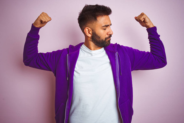 孤立したピンクの背景の上に立つ紫色のスウェットシャツを着た若いインド人男性は、腕の筋肉を誇りに思って笑顔。フィットネスコンセプト. - 写真・画像