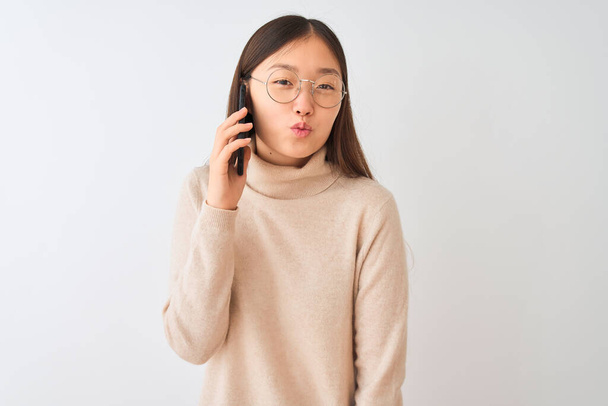 Nuori kiinalainen nainen puhuu älypuhelimella yli eristetty valkoinen tausta katselee kameraa puhaltaa suudelma ilmassa on ihana ja seksikäs. Rakkauden ilme
. - Valokuva, kuva