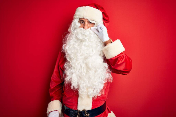 Ein gutaussehender Mann mittleren Alters im Weihnachtsmannkostüm, der vor isoliertem roten Hintergrund steht und die Lippen als Reißverschluss mit den Fingern geschlossen hält. Geheimniskrämerei und Schweigen, Tabuthema - Foto, Bild