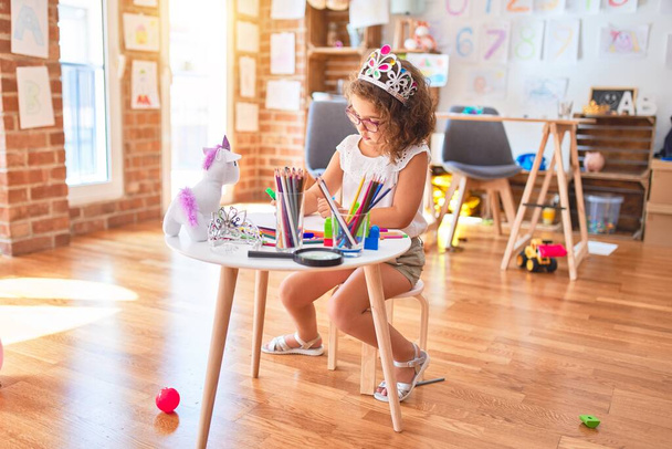 Όμορφο νήπιο φορώντας γυαλιά και πριγκίπισσα στέμμα κάθεται σχέδιο χρησιμοποιώντας χαρτί και μαρκαδόρο στυλό στο νηπιαγωγείο - Φωτογραφία, εικόνα