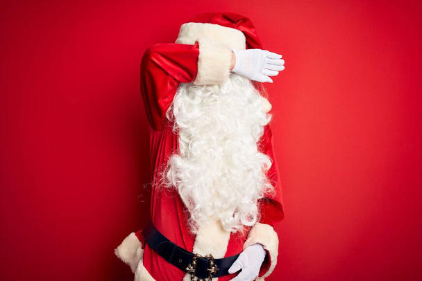 Orta yaşlı yakışıklı adam, Noel Baba kostümü giymiş, izole edilmiş kırmızı arka plan üzerinde duruyor, gözleri kolla kaplıyor, ciddi ve üzgün görünüyor. Görme, saklama ve reddetme konsepti - Fotoğraf, Görsel