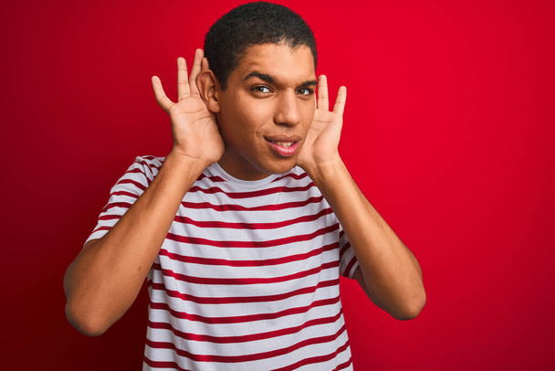 Young handsome arab man wearing striped t-shirt over isolated red background Trying to hear both hands on ear gesture, curious for gossip. Hearing problem, deaf - Photo, Image