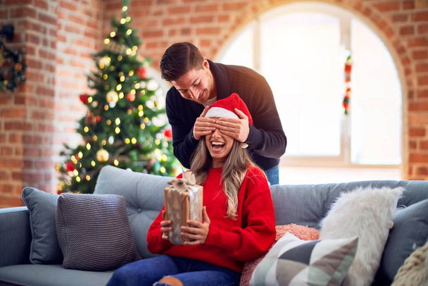 Молодая красивая пара улыбается счастливой и уверенной. Мужчина, закрывающий глаза женщины, окружает её подарком вокруг рождественской елки дома
 - Фото, изображение