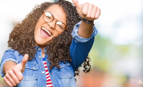 Young beautiful woman with curly hair wearing glasses approving doing positive gesture with hand, thumbs up smiling and happy for success. Looking at the camera, winner gesture. - Photo, Image