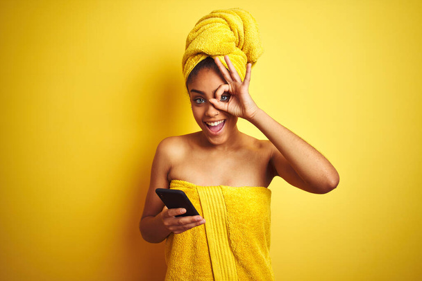 シャワー後のタオルを身に着けているアフロ女性は指を通して見て目に手でOKサインを行う幸せな顔の笑顔で隔離された黄色の背景にスマートフォンを使用して - 写真・画像