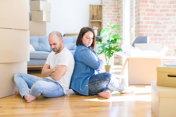 段ボール箱の周りの床の上に座っている若いカップルは、新しい家の懐疑的で緊張し、両腕を組んで顔に不承認式に移動します。負の人. - 写真・画像