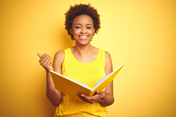 Afrikanische Amerikanerin liest ein Buch über gelbem, isoliertem Hintergrund und zeigt mit dem Daumen nach oben zur Seite mit glücklichem Lächeln - Foto, Bild