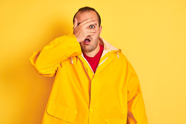 Νεαρός άνδρας φορώντας παλτό βροχής στέκεται πάνω από απομονωμένο κίτρινο φόντο κρυφοκοιτάζει σε σοκ καλύπτει το πρόσωπο και τα μάτια με το χέρι, κοιτάζοντας μέσα από τα δάχτυλα με αμηχανία έκφραση. - Φωτογραφία, εικόνα